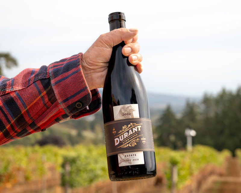 A hand wearing a red and black plaid shirt holds up a bottle of Durant Vineyard Estate Pinot Noir against a blurred vineyard background.