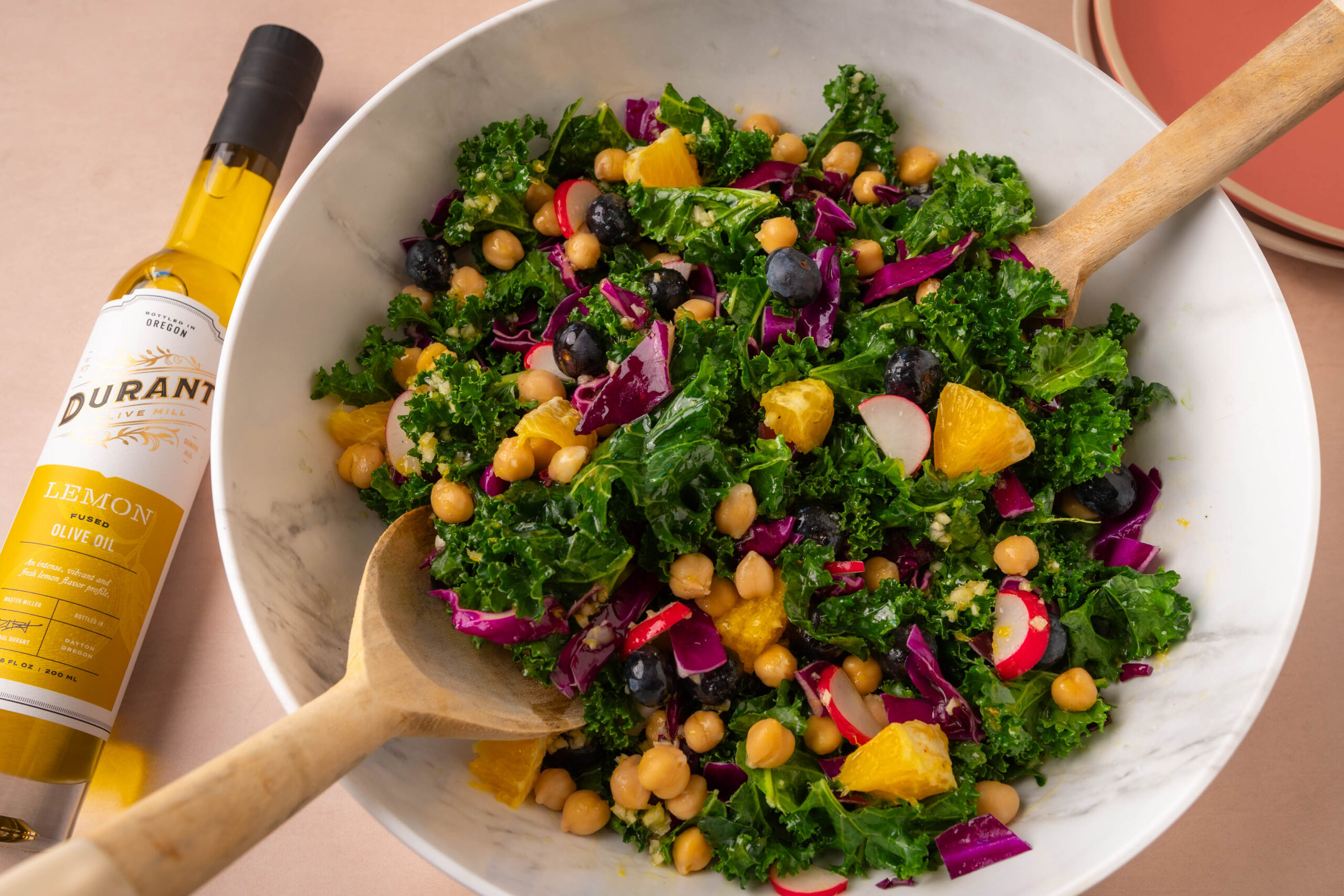 Picture of Durant Kale Rainbow salad in a white bowl. The green kale salad is mixed with chickpeas, red shredded cabbage slices, orange slices and blueberries throughout. There is a bottle of Durant Lemon Fused Olive Oil to the left of the bowl, laying down on the table, and wood salad mixing spoons in the bowl.